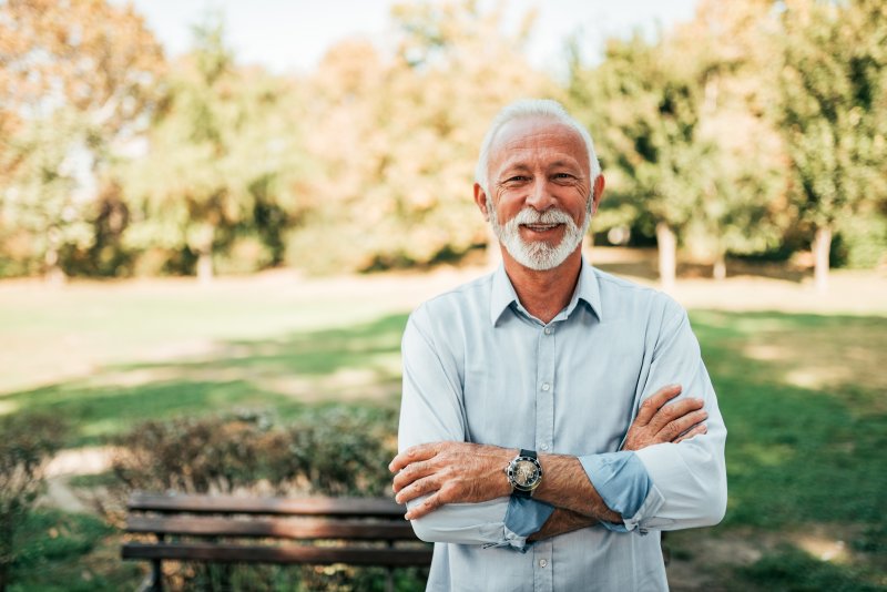 an older man standing outside with his arms crossed and smiling after receiving his dental implants 