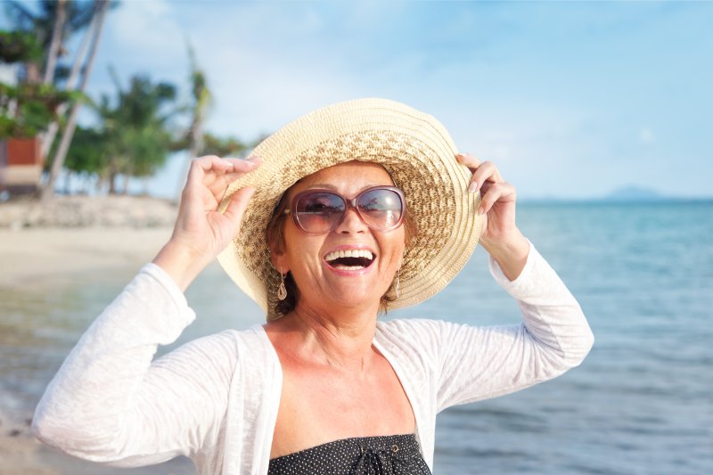 an older woman holding onto her hat while standing in the ocean and smiling after receiving dental implants in Jacksonville