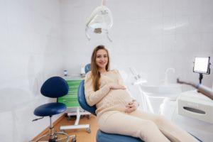 a pregnant person sitting in a dental treatment chair and smiling while holding their belly